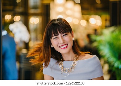 Portrait Of An Older Asian Woman Standing And Posing For Her Head Shot Standing In The Doorway Of A Warmly Lit Room During The Day. She Is Smiling Happily. 