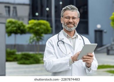 Portrait of an older gray-haired male doctor standing outside the clinic in a white coat with a stethoscope. He holds a tablet in his hands, looks at the camera, smiles. - Powered by Shutterstock