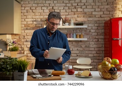 Portrait Of An Older Man. Older Man Having Breakfast In Kitchen, Reading Morning News On Tablet Computer.