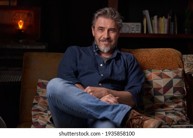Portrait of older man at home in dark room sitting on couch. Happy smile, grey hair, bearded, . mature age, middle age, mid adult man in 50s.