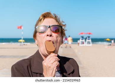 Portrait Of An Older Woman, Eating An Ice Cream, With Fun And Colorful Sunglasses, On A Sunny Day At The Beach.