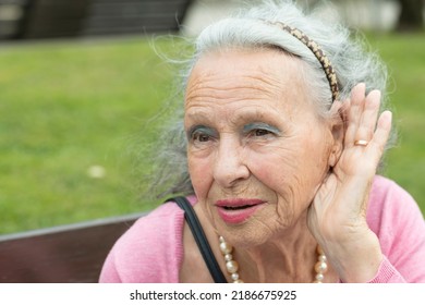 Portrait Of An Older Woman Who Is Hard Of Hearing And Deaf. Woman In The Park With Her Hand On Her Ear Due To Hearing Problems And Deafness. Concept Of Hearing Loss In The Elderly. Hearing Loss.