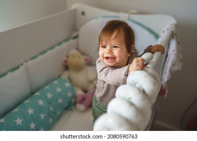 Portrait Of One Happy Baby Girl 7 Months Old Standing In A Crib With Toys In A Bright Children's Room After Sleeping And Looking Around Copy Space