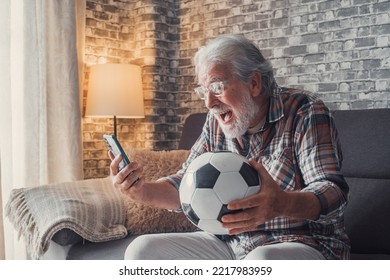 Portrait Of One Happy Old Man Celebrating Victory Of His Love Team Of Football Or Soccer On The Sofa. Senior Watching Match On His Phone Winning A Football Bet.