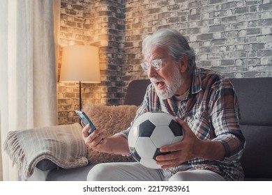 Portrait Of One Happy Old Man Celebrating Victory Of His Love Team Of Football Or Soccer On The Sofa. Senior Watching Match On His Phone Winning A Football Bet.