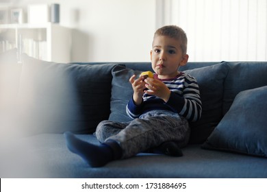 Portrait Of One Small Three Years Old Caucasian Boy Little Child Kid Sitting On The Sofa Bed At Home Holding Chocolate Candy In Hand Eating While Watching Tv Front View