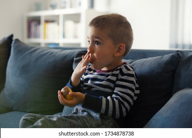 Portrait Of One Small Three Years Old Caucasian Boy Little Child Kid Sitting On The Sofa Bed At Home Holding Chocolate Candy In Hand Eating While Watching Tv Lick Finger In Mouth Side View