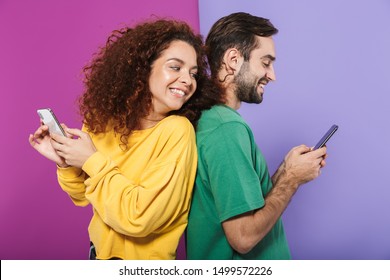 Portrait of optimistic caucasian couple in colorful clothing smiling and holding cellphones standing back to back isolated over violet background