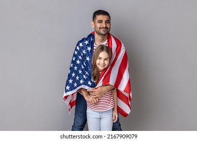Portrait of optimistic father and daughter standing wrapped in big flag of United States of America together, celebrating national holiday. Indoor studio shot isolated on gray background.