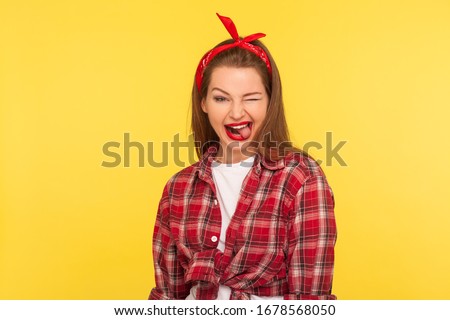 Portrait of optimistic pinup girl in checkered shirt and headband winking to camera with crazy naughty expression, showing tongue out, making faces blinking eye. retro 50's style, studio shot isolated