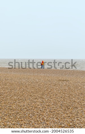 Portrait orientation shot of two distant people hugging on an empty beach, colourful jacket clothing, windy cold weather, stones on beach, sea in background, dull looking sky, no faces