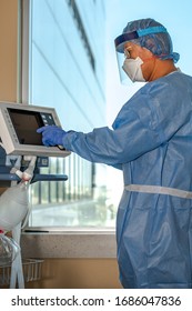 A Portrait Oriented Photo Of A Respiratory Therapist In Full Personal Protective Equipment Makes Changes To A Ventilator In An Intensive Care Or Emergency Room With Window In The Background.