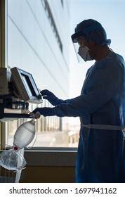 A Portrait Oriented Photograph Of A Respiratory Therapist Silhouetted Against Window Makes Changes To A Ventilator In Full Protective Gear For A Patient With An Influenza Like Illness Such As Covid-19