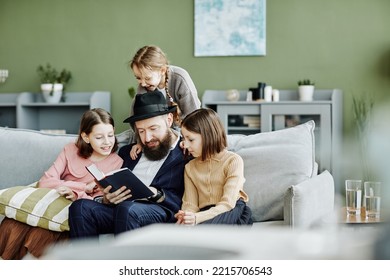 Portrait of orthodox Jewish family with father reading book to three children in modern home interior, copy space