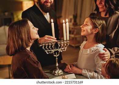 Portrait Of Orthodox Jewish Family Lighting Menorah Candle Together During Hanukkah Celebration With Focus On Two Girls Smiling