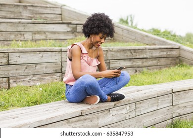 Portrait Outdoors Of A Beautiful Young Afro American Woman At Sunset Using Mobile Phone And Smiling. Wood Background. Lifestyle.Casual Clothing. Sitting On Wood Stairs And Smiling.