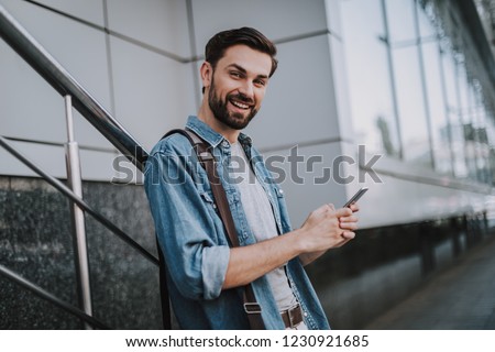 Portrait of outgoing bearded male typing in mobile while looking at camera outdoor