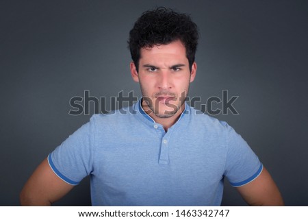 Portrait of outraged young man with oval face, wearing blue T-shirt frowning his eyebrows being displeased with something. Scowling pretty female isolated over grey.