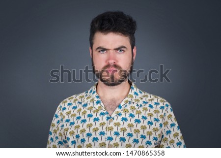 Portrait of outraged young man with oval face, blue eyes wearing palm shirt frowning his eyebrows being displeased with something. Scowling pretty female isolated over grey.