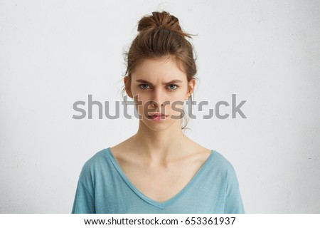 Portrait of outraged young woman with oval face, blue eyes and hair bun wearing blue casual sweater frowning her eyebrows being displeased with something. Scowling pretty female isolated over white