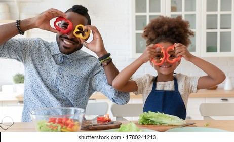 Portrait Of Overjoyed African American Young Dad And Little Daughter Make Funny Faces With Vegetable Cooking Together, Happy Biracial Father And Small Girl Child Have Fun Preparing Food In Kitchen