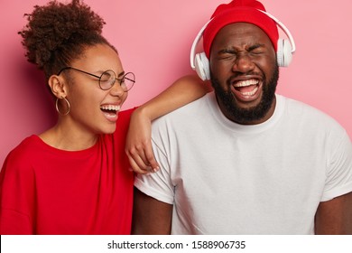 Portrait Of Overjoyed Dark Skinned Female And Male Laugh With Fun, Have Happy Moods, Guy Listens Audio Track In Headphones, Stand Closely Over Pink Wall. Cheerful Afro Couple Spend Time Together