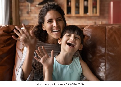 Portrait of overjoyed Hispanic young mom and little daughter wave greet at camera talk on video call at home. Smiling Latino mother and small 8s girl child have webcam digital virtual online event.