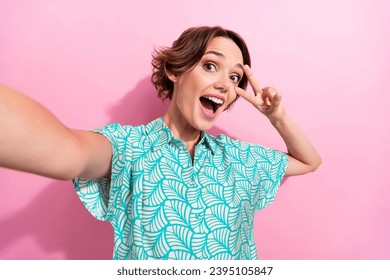 Portrait of overjoyed nice girl make selfie demonstrate v-sign near eye isolated on pink color background