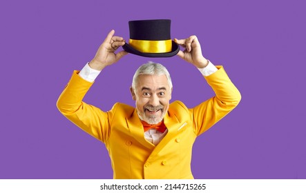 Portrait Of Overjoyed Old Man In Jacket And Hat Isolated On Purple Studio Background Show Tricks And Jokes. Funny Smiling Elderly Male In Suit Have Fun Entertain Audience. Entertainment Concept.