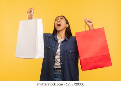 Portrait Of Overjoyed Shopper Girl In Stylish Denim Shirt Raising Bags And Shouting From Happiness, Amazed Thrilled By Shopping In Fashion Store, Thrift Sale. Studio Shot Isolated On Yellow Background