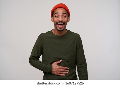 Portrait of overjoyed young bearded dark skinned guy looking at camera with wide eyes opened and holding hand on his belly while laughing happily, isolated over white background