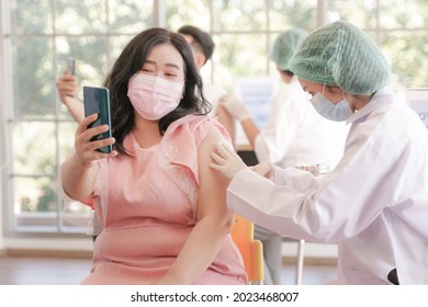 Portrait Of Overweight Asian Woman Selfie By Phone At While Getting Covid Vaccine In Clinic Or Hospital, With Hand Nurse Injecting Vaccine To Get Immunity From Virus. People Wearing Protective Mask.