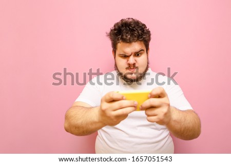 Portrait of an overweight young overweight man playing mobile games on a sad-faced smartphone. Fat gamer guy plays online games on his phone and looks intently at the camera wearing a white T-shirt