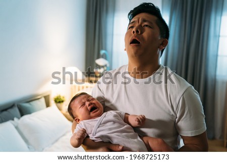 portrait overwhelmed asian father is holding his crying baby daughter and looking upward in despair at midnight in the bedroom at home.