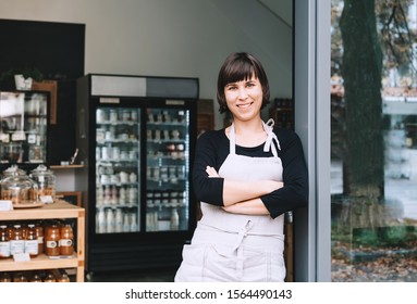 Portrait of owner of sustainable small local business. Shopkeeper of zero waste shop standing on interior background of shop. Smiling young woman in apron welcoming at entrance of plastic free store - Powered by Shutterstock