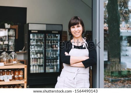 Portrait of owner of sustainable small local business. Shopkeeper of zero waste shop standing on interior background of shop. Smiling young woman in apron welcoming at entrance of plastic free store