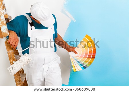 portrait of painter man with paint roller and wooden vintage ladder showing a color palette, on half painted wall
