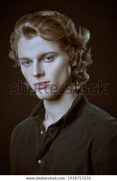Portrait of a pale handsome young man with wavy\
blond hair in dark shirt looking at the camera. Black background\
with copy space. Men\'s\
beauty.