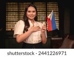 Portrait of a patriotic young woman holding a Philippine flag, dressed in elegant traditional Filipino attire. Putting his hand in her heart, showing love to country.