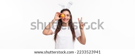Portrait of peaceful and cheerful young cute female in white t-shirt, showint peace sign and hold colored egg over eye, smiling happy, enjoying traditional holiday celebration, Easter day.