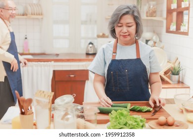 Portrait Of Pensioner Mature Woman Chopping Cucumber For Vegetable Salad Dinner. Senior Retired Influencer Woman Making Healthy Food In The Kitchen At Home. Elder People Lifestyle Concept.