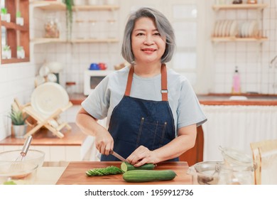 Portrait Of Pensioner Mature Woman Chopping Cucumber For Vegetable Salad Dinner. Senior Retired Influencer Woman Making Healthy Food In The Kitchen At Home. Elder People Lifestyle Concept.