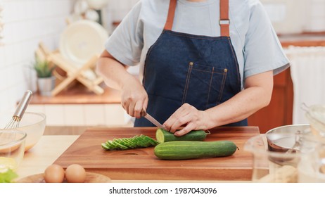 Portrait Of Pensioner Mature Woman Chopping Cucumber For Vegetable Salad Dinner. Senior Retired Influencer Woman Making Healthy Food In The Kitchen At Home. Elder People Lifestyle Concept.