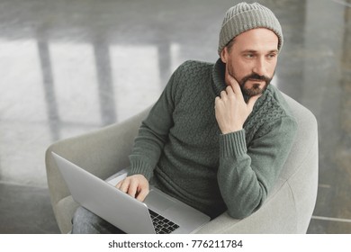 Portrait of pensive bearded middle aged male writer wears knitted sweater and hat, sits in comfortable armchair, keyboards on laptop new chapter of book, tries to image happy end. Businessman indoor