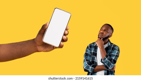 Portrait Of Pensive Black Guy Thinking And Looking At Huge White Blank Cell Screen, Giant Hand Holding Huge Smartphone, Showing Device Gadget With Empty Free Copy Space For Mock Up, Yellow Orange Wall