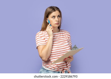 Portrait of pensive blond woman wearing striped T-shirt taking notes with pen on clipboard, standing deep in thoughts, thinking. Indoor studio shot isolated on purple background.