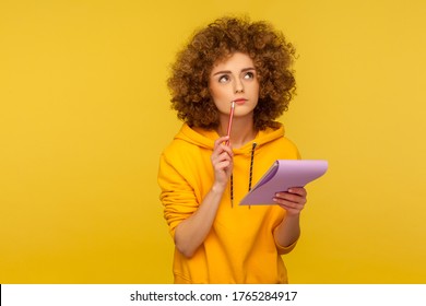 Portrait of pensive curly-haired hipster woman in urban style hoodie thinking over smart idea, holding pencil and notebook to write plans, to-do list. indoor studio shot isolated on yellow background - Powered by Shutterstock