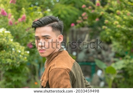 Portrait of a pensive Gen Z man in a cool jacket posing at a scenic garden resort.