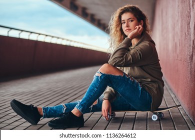 Portrait Of A Pensive Girl In A Hoodie And Ripped Jeans Sitting On A Skateboard At Bridge Footway.