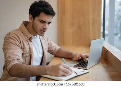 Portrait Of Pensive Indian Businessman Working In Modern Office. Handsome Man Freelancer Using Laptop Computer, Typing, Taking Notes On Notebook. University Student Studying, Exam Preparation